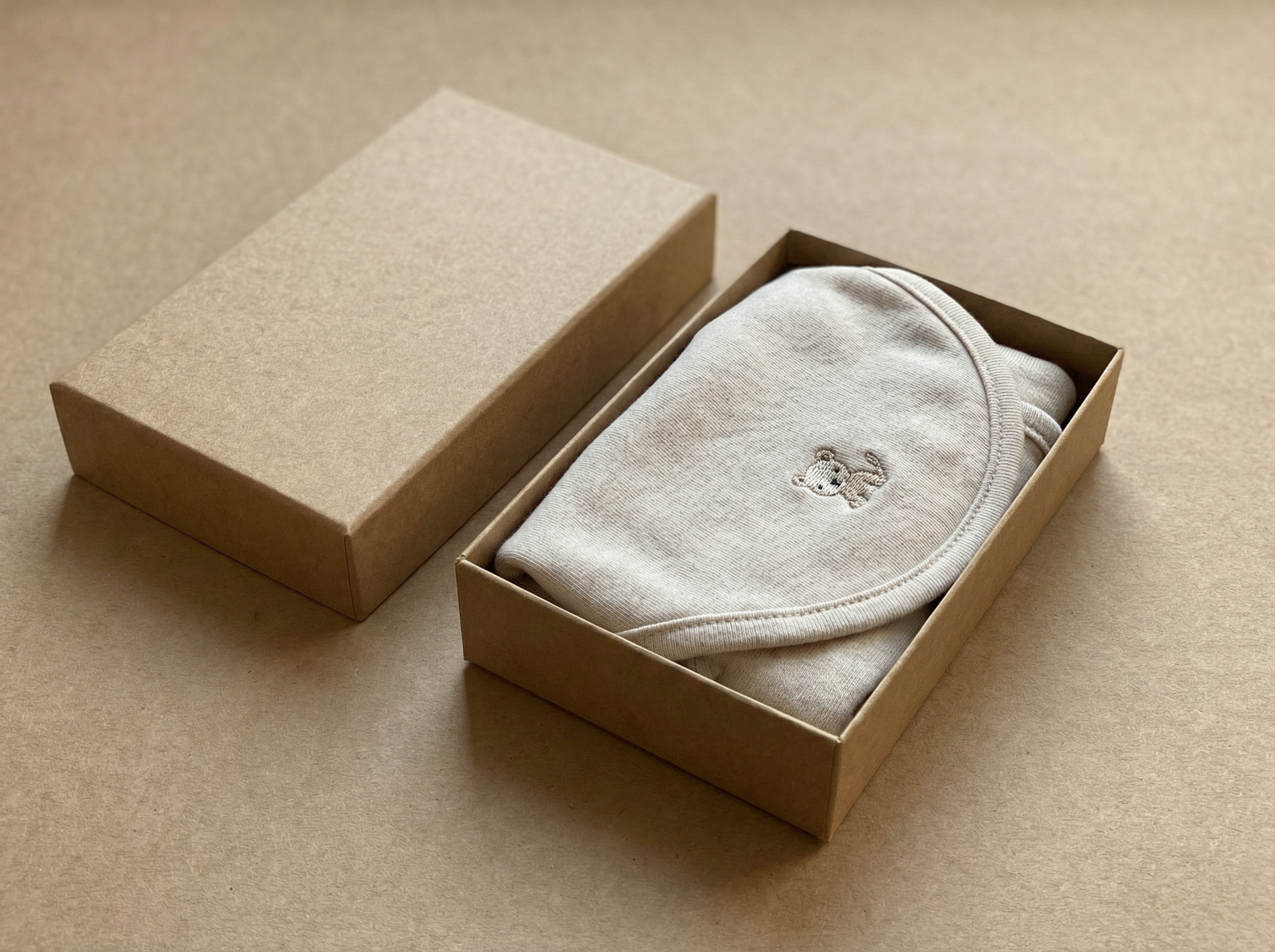 Boîte en carton ouverte contenant un vêtement beige plié avec broderie d'un petit ours, couvercle posé à côté.