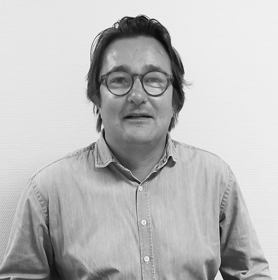 Homme portant des lunettes rondes et une chemise en denim, posé devant un mur texturé, regardant l'appareil.