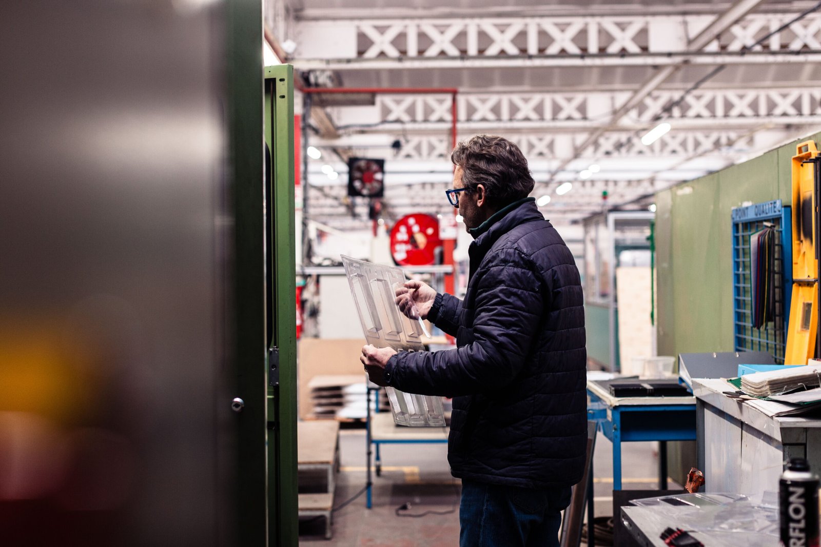 Ouvrier en veste matelassée examinant une pièce plastique transparente dans un atelier industriel.