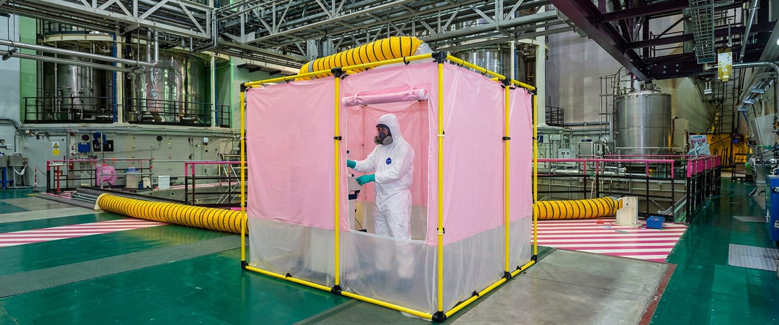 Technicien en combinaison intégrale et masque travaillant dans une cabine rose portable dans une grande enceinte industrielle avec tuyaux jaunes d'aération.
