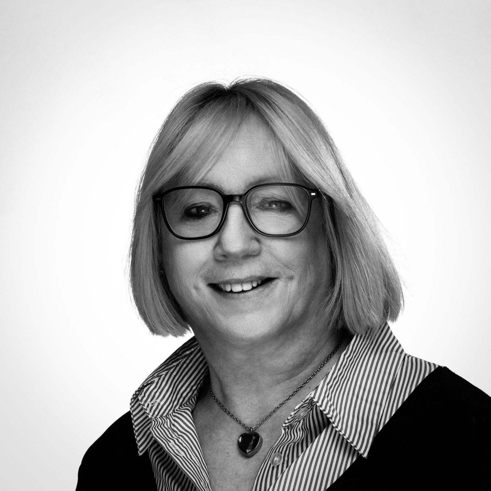 Femme souriante aux cheveux mi-longs, lunettes épaisses et collier cœur, chemise rayée et fond uni.